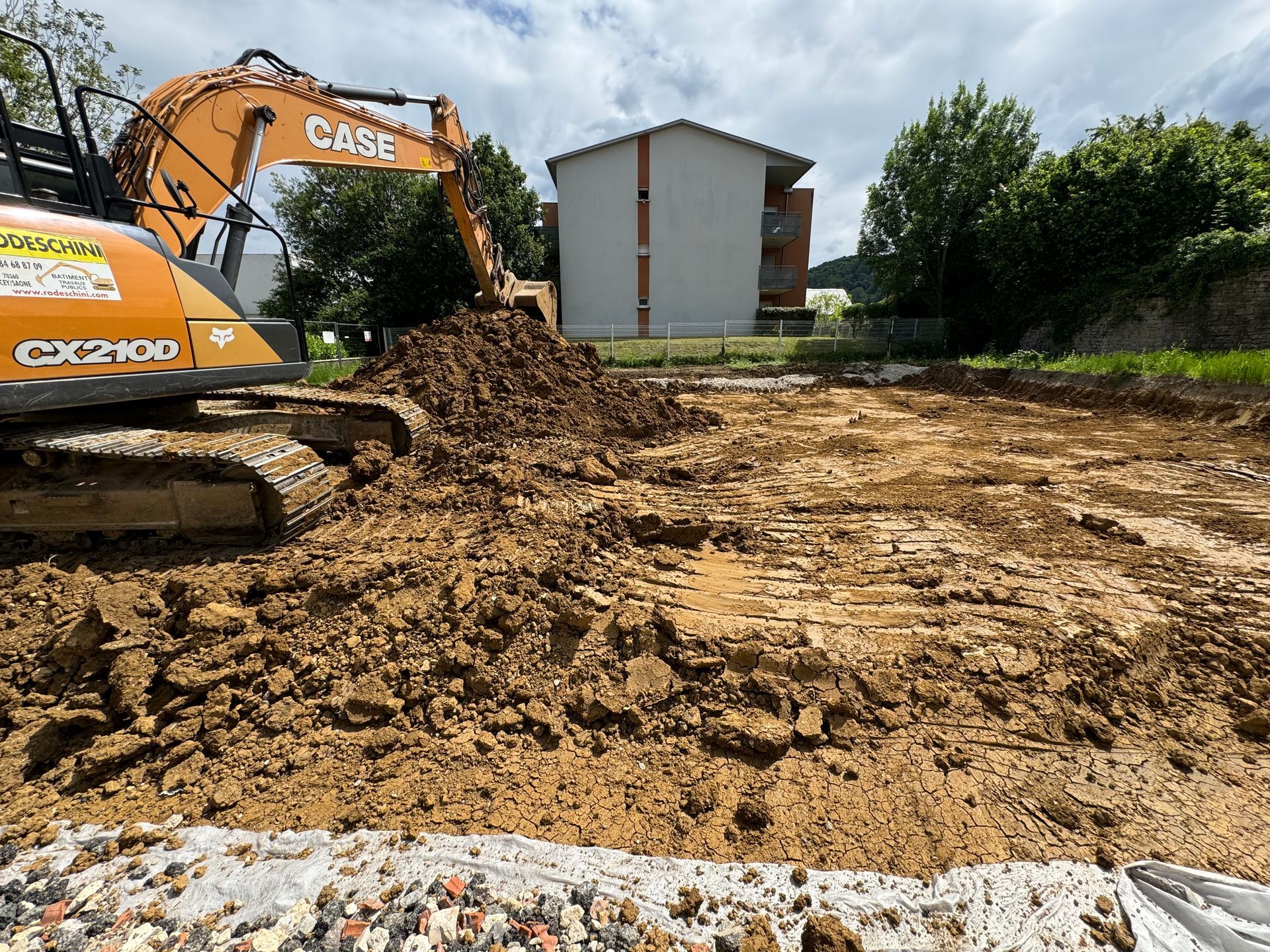 Une excavatrice déplace de la terre sur un chantier de construction ; un bâtiment et des arbres sont en arrière-plan