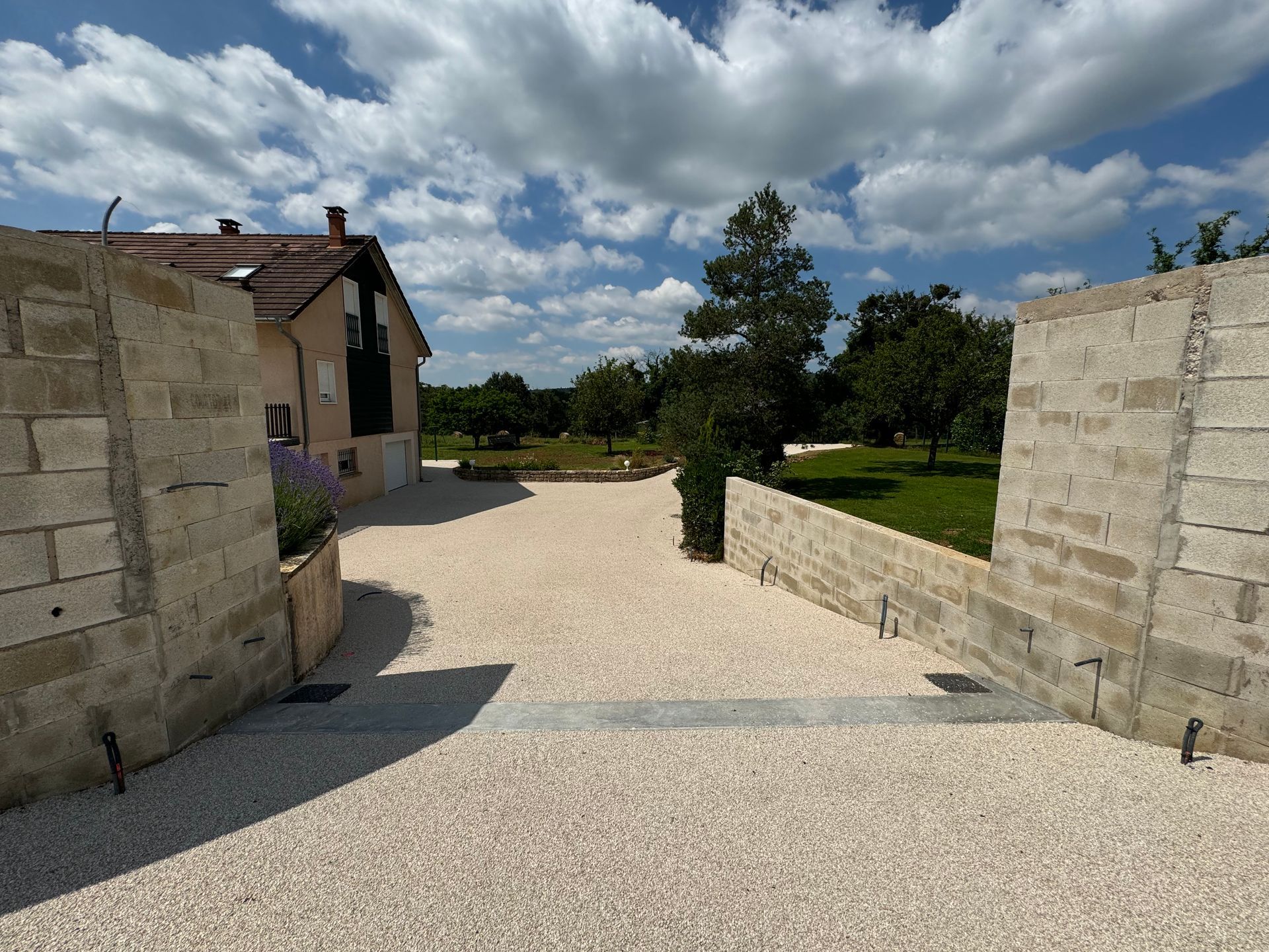Allée de maison avec gravier, bordée de murets, menant à une cour et des arbres