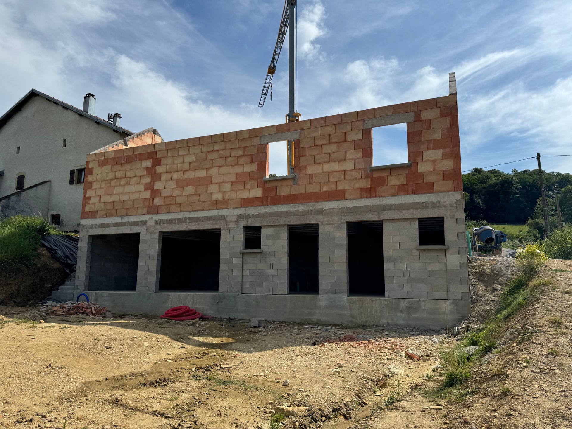 Maison en construction : murs en briques et parpaings, fenêtres découpées, grue au-dessus