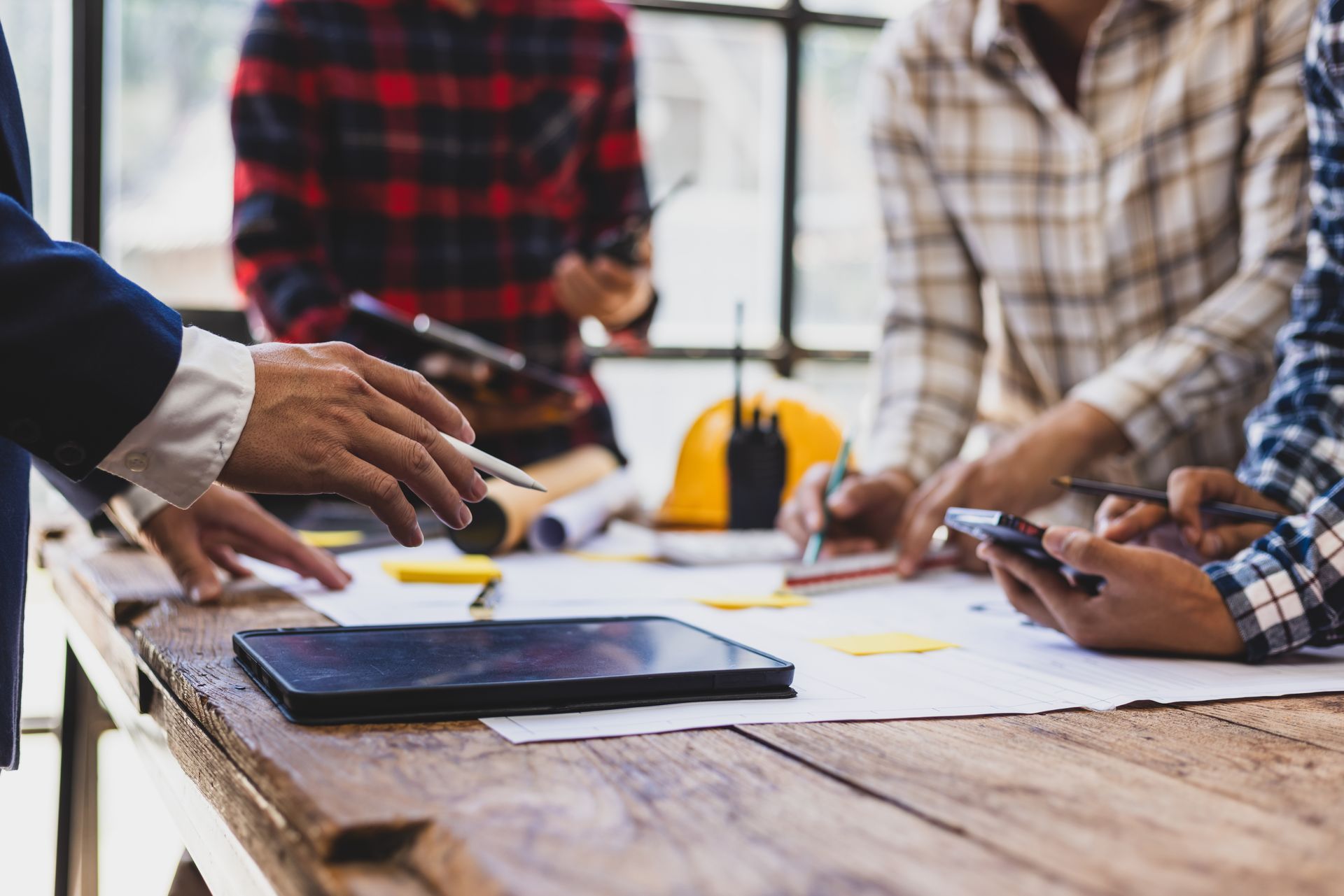 Des personnes collaborent autour d'une table en bois avec des papiers, une tablette et des écouteurs jaunes dans un environnement de bureau lumineux.