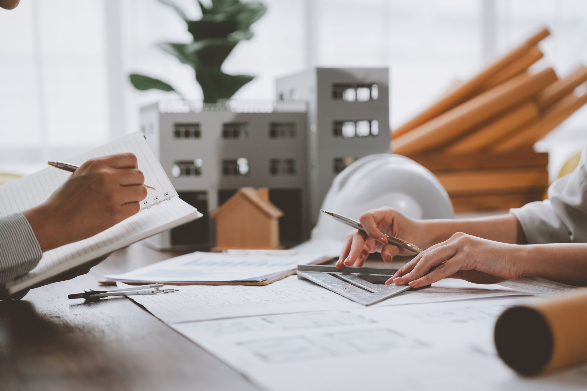 Des mains examinent des plans architecturaux et des maquettes de bâtiments sur une table dans un bureau lumineux.