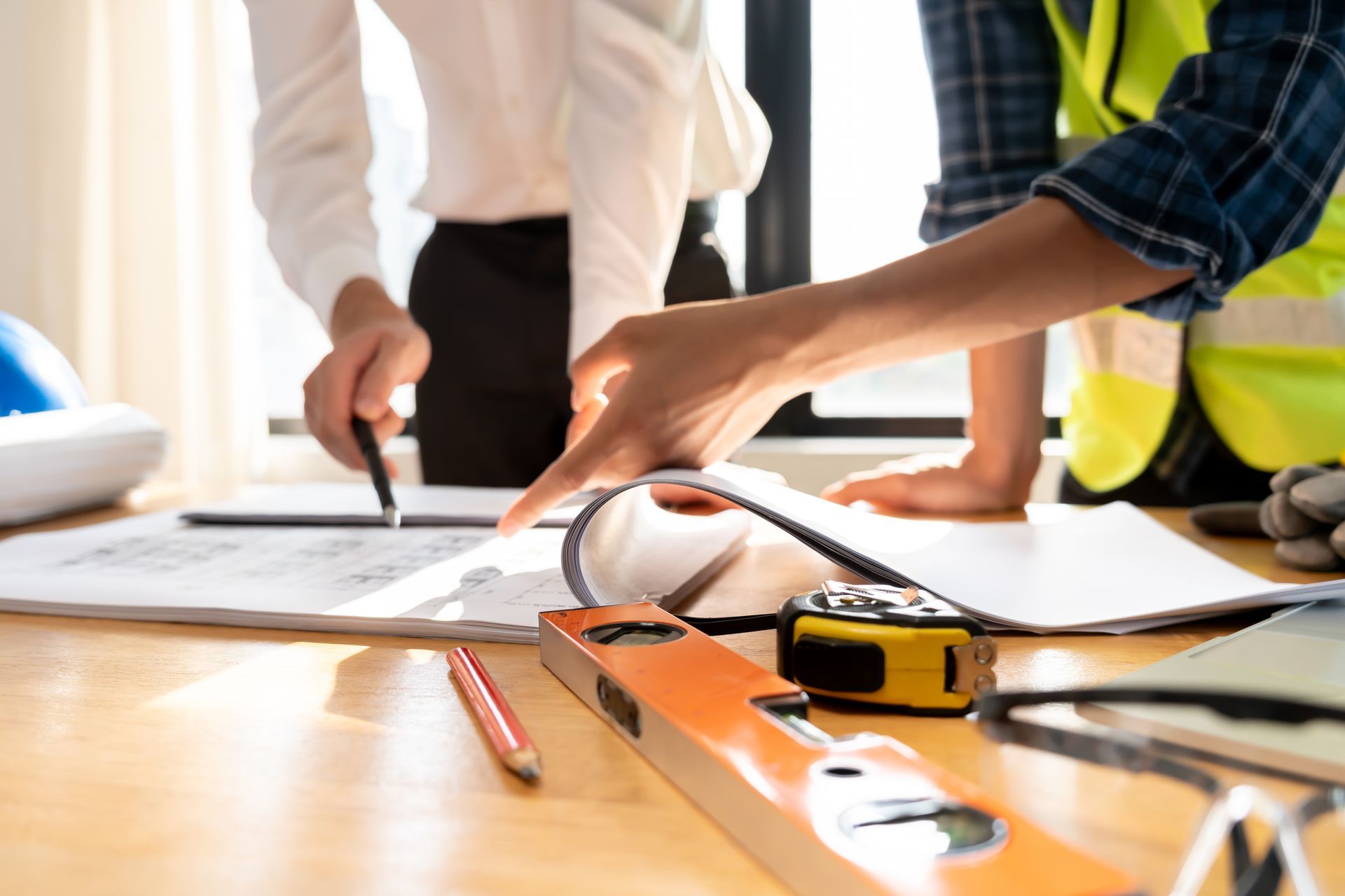 Des personnes examinant des plans sur un bureau, munies d'un crayon, d'un mètre ruban et d'un casque de chantier, dans un environnement de construction.
