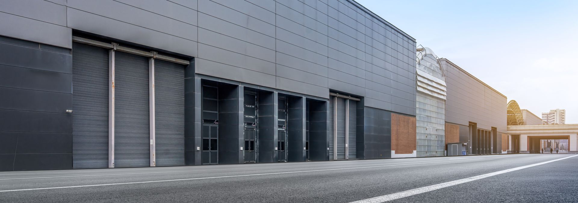 Rangée d'entrepôts industriels aux façades métalliques grises et aux portes de chargement fermées, le long d'une rue déserte au lever du soleil