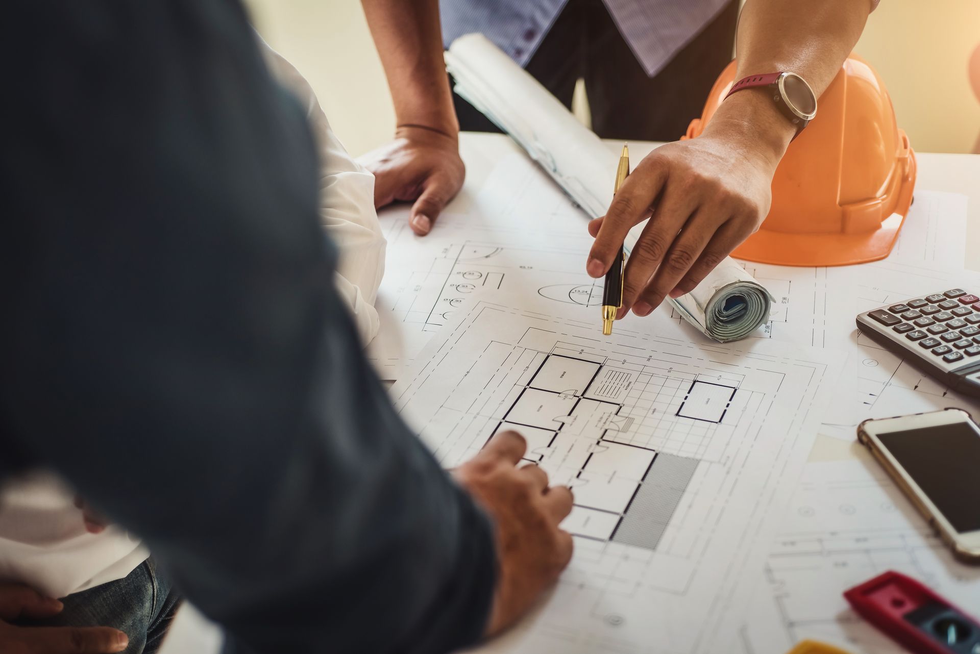 Des mains examinant un plan de construction, un casque de chantier, un mètre ruban et une calculatrice posés sur la table.