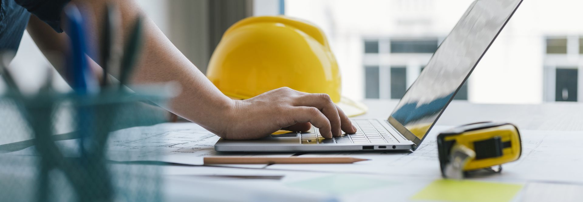 Une main tapant sur un ordinateur portable à côté d'un casque de chantier jaune dans un bureau lumineux