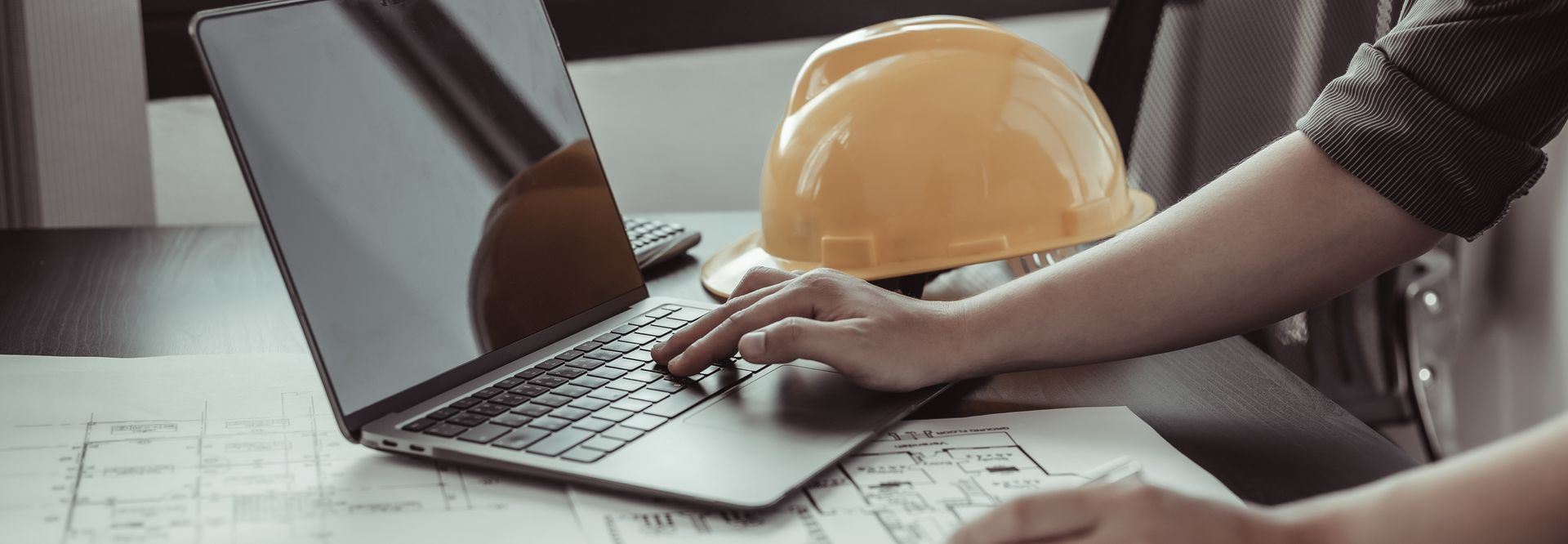 Une personne utilise un ordinateur portable à côté d'un casque de chantier jaune posé sur un bureau avec des plans de construction.