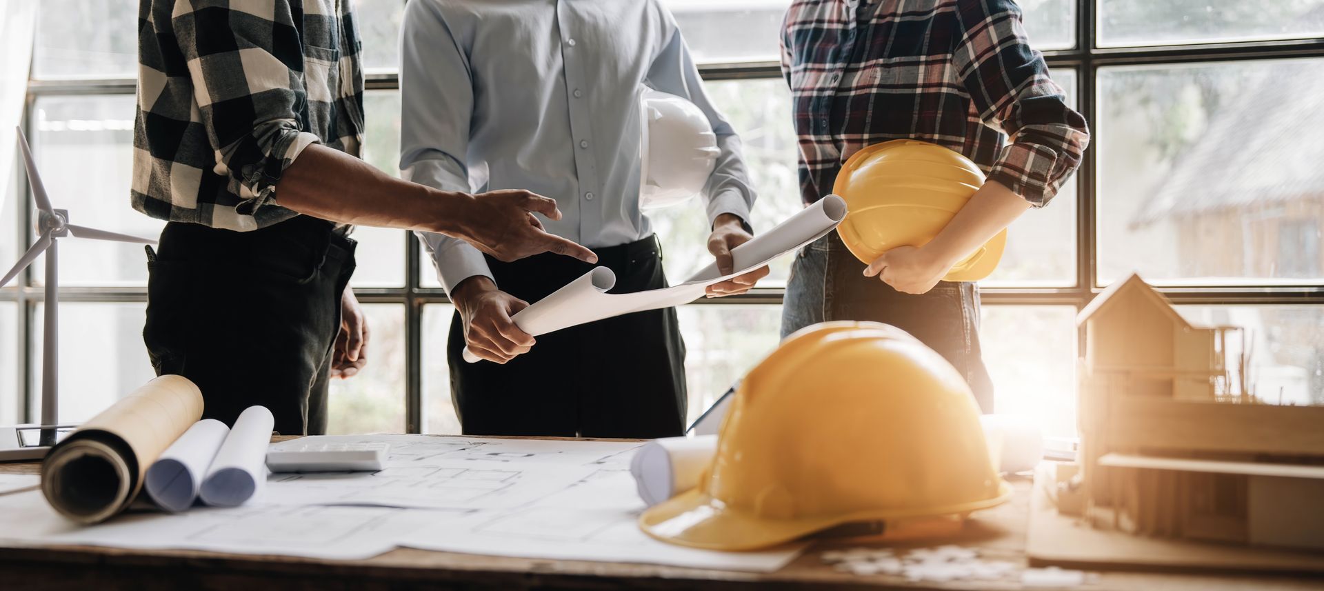 Des ingénieurs en réunion examinent des plans à côté de casques de chantier jaunes dans un bureau lumineux.