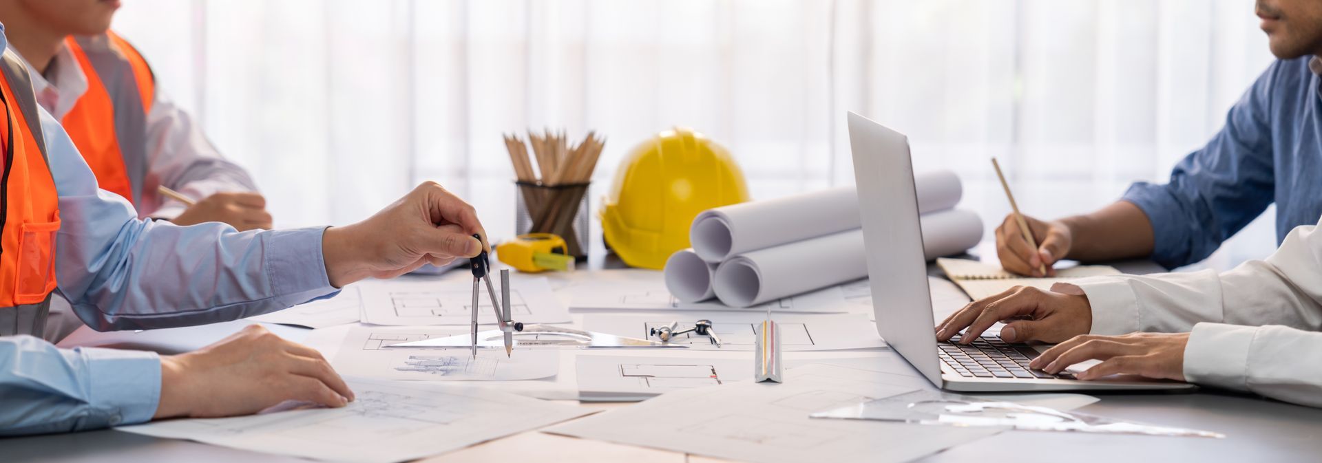 Une équipe de collègues examine des plans autour d'une table, avec des plans, un ordinateur portable et un casque de chantier, dans un bureau lumineux.