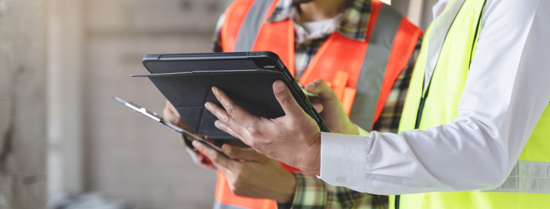 Ouvrier du bâtiment tenant une tablette et des documents, portant un gilet de sécurité et un casque de chantier.
