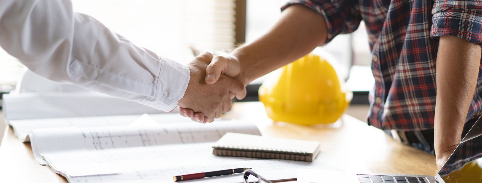 Deux personnes se serrent la main au-dessus de plans sur un bureau, avec un casque de chantier jaune en arrière-plan.
