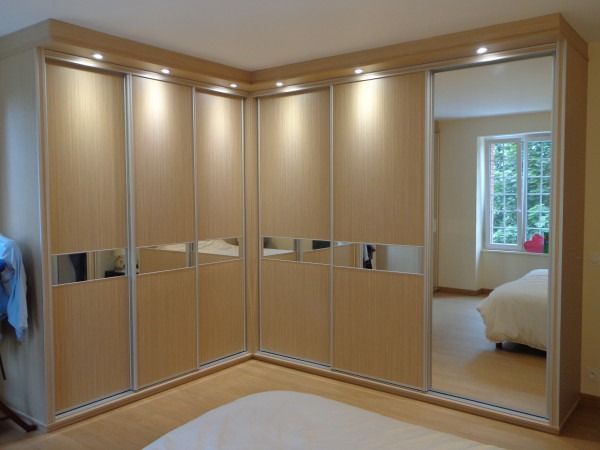 Armoire d'angle avec portes en bois clair, garnitures argentées et panneaux miroir, dans une chambre.