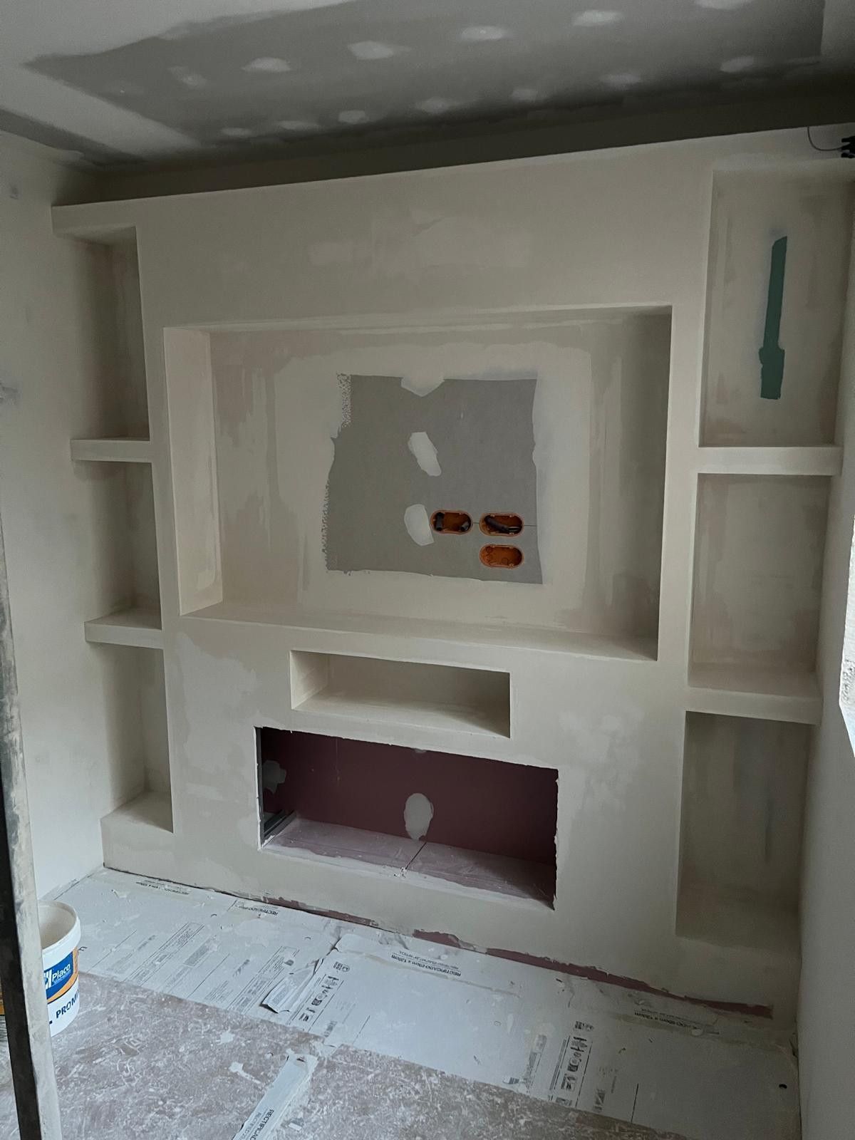 Mueble empotrado blanco con estantes, nicho central y aberturas recortadas en una habitación sin terminar con revestimiento protector de suelo.