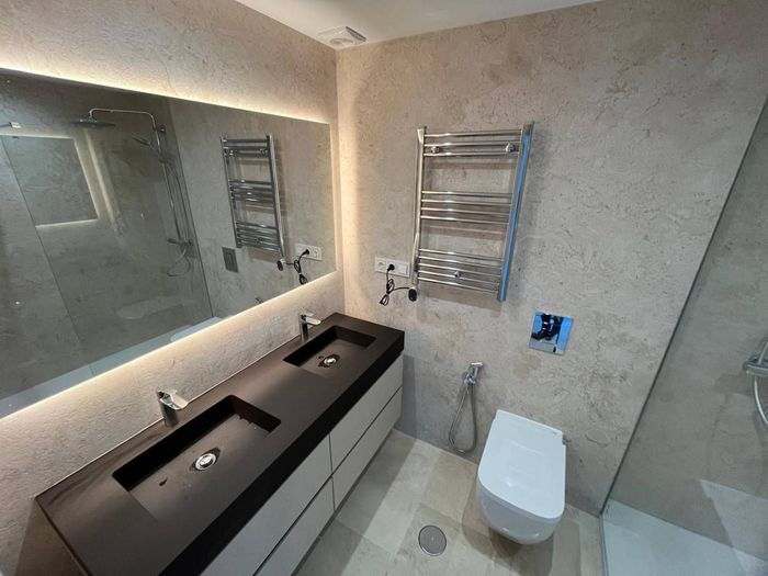Baño moderno con tocador de doble lavabo, inodoro, espejo grande, toalleros eléctricos y ducha independiente.