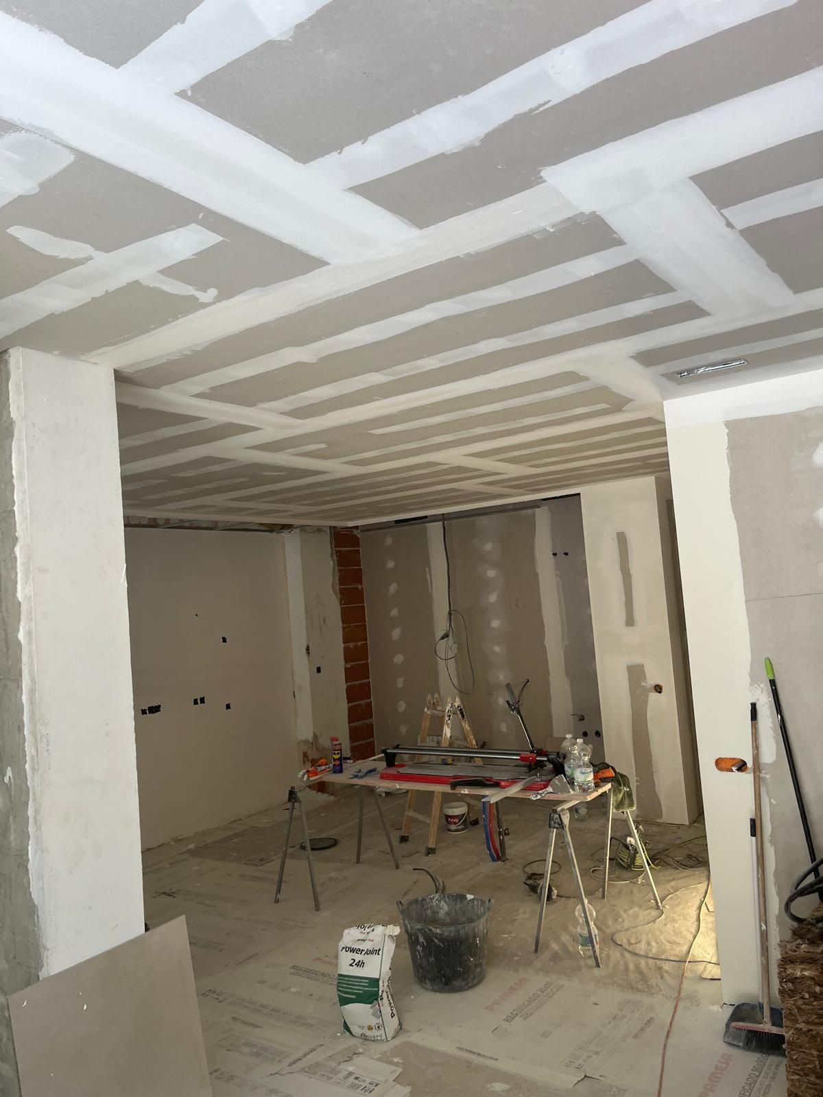 Habitación de reforma con paneles de yeso, herramientas y materiales de construcción sobre un suelo y techo parcialmente terminados.