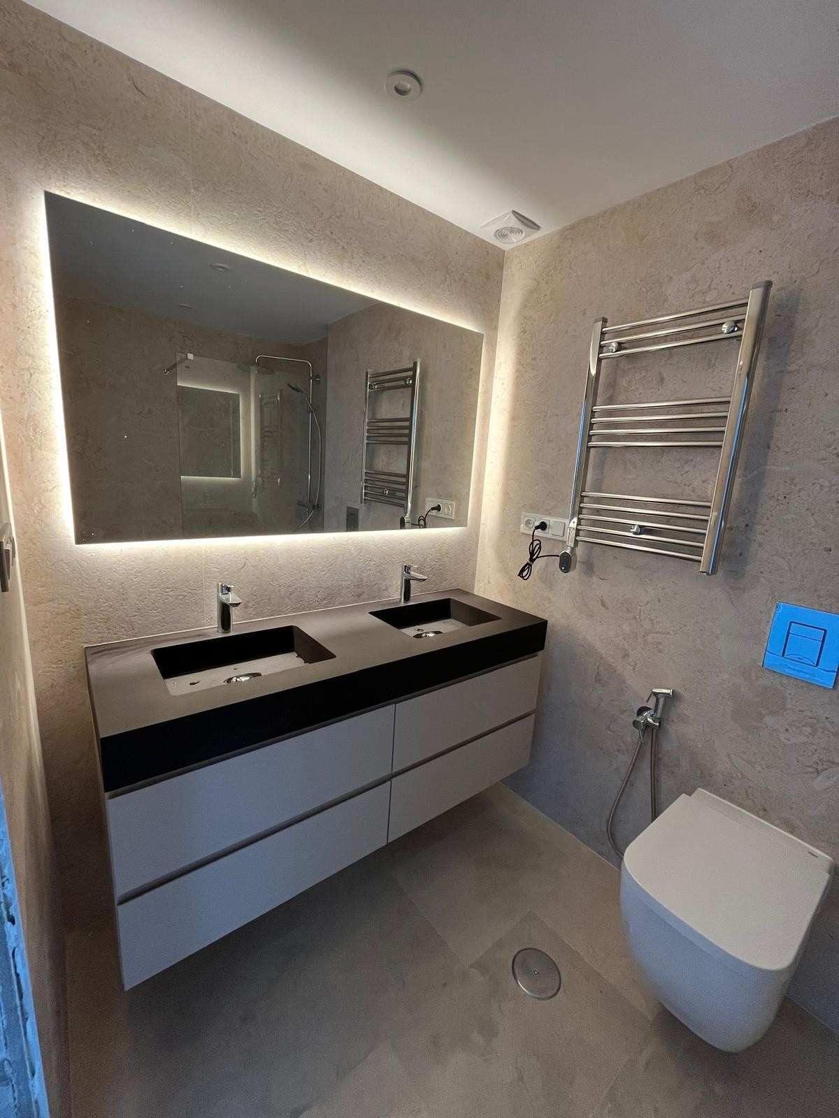 Baño moderno con lavabo doble, espejo grande, toallero eléctrico e inodoro suspendido bajo iluminación tenue.