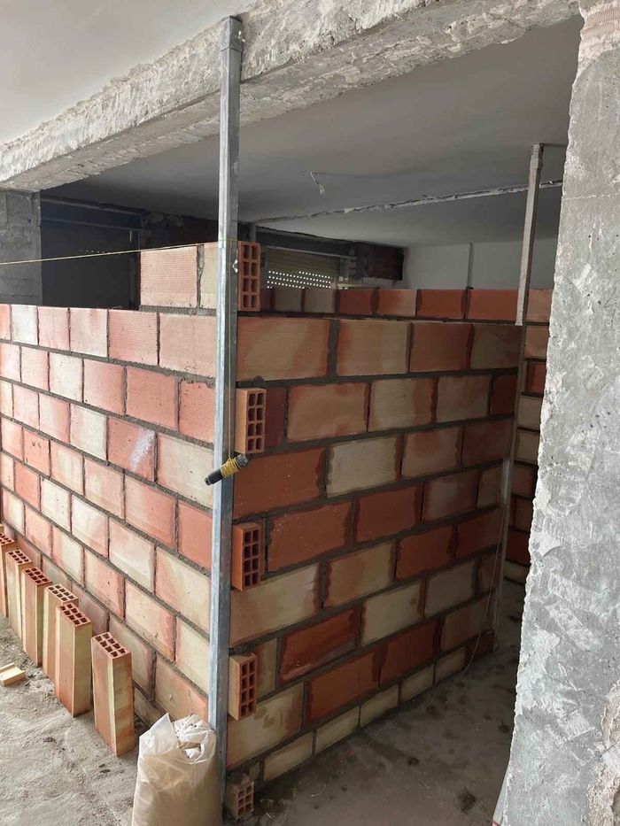 Muro de ladrillo parcialmente construido, sostenido por un puntal metálico vertical dentro de una estructura de hormigón.