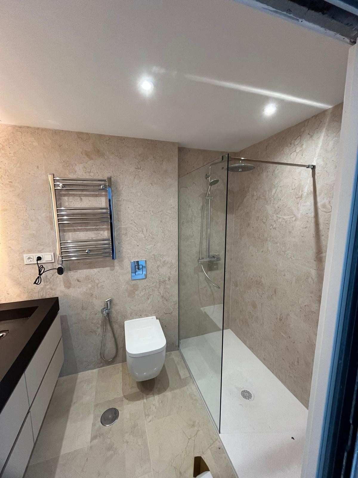 Baño moderno con inodoro, ducha de cristal, paredes de azulejos beige y toallero eléctrico.