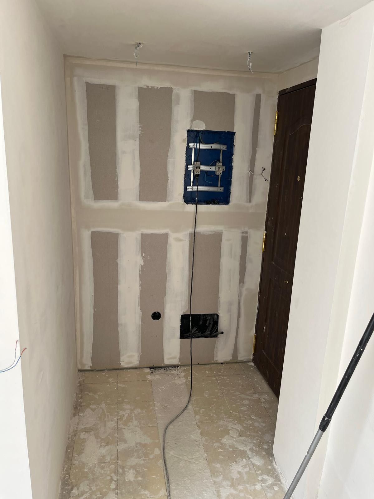 Habitación estrecha sin terminar con parches de paneles de yeso, cableado expuesto y una pequeña caja eléctrica azul en la pared del fondo.