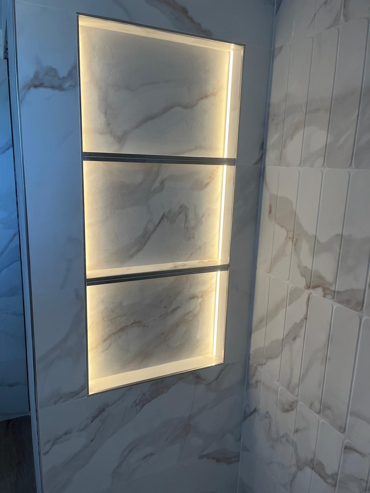 Nicho de ducha empotrado iluminado con azulejos con estampado de mármol en un baño moderno.