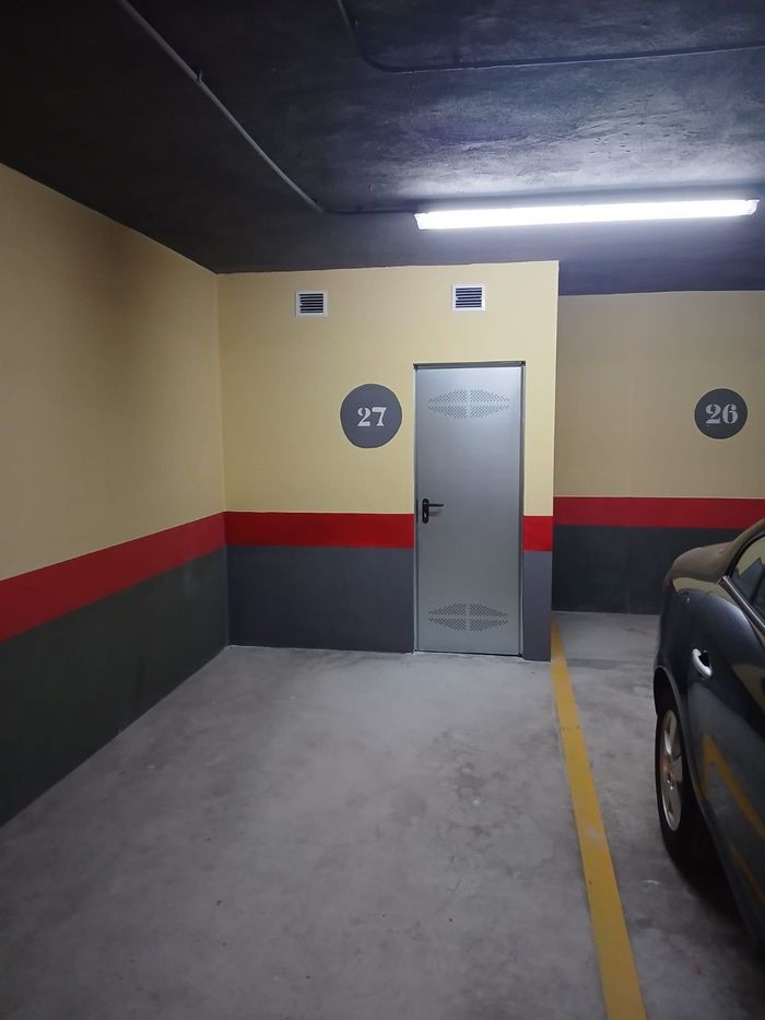 Pasillo de un aparcamiento subterráneo con una puerta cerrada, paredes rojas y negras, y un coche aparcado a la derecha.