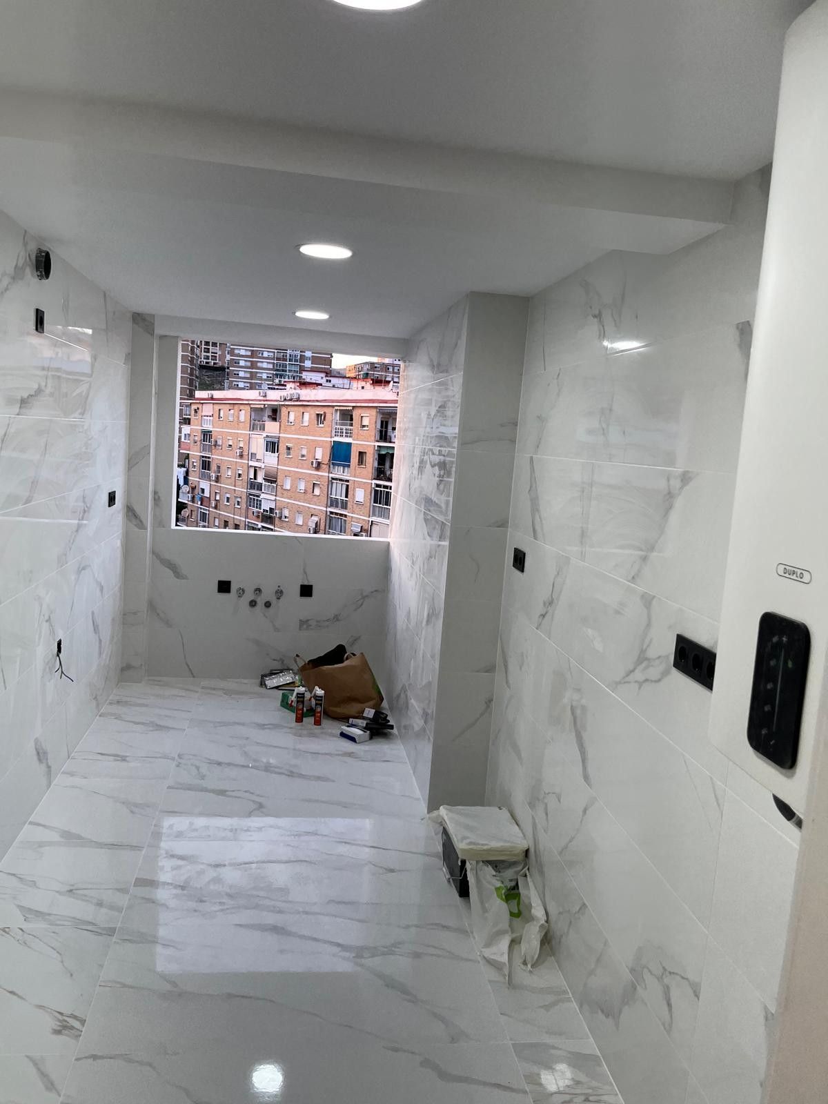 Pasillo con baldosas blancas en obras, con herramientas en el suelo y una ventana con vistas a edificios de ladrillo.