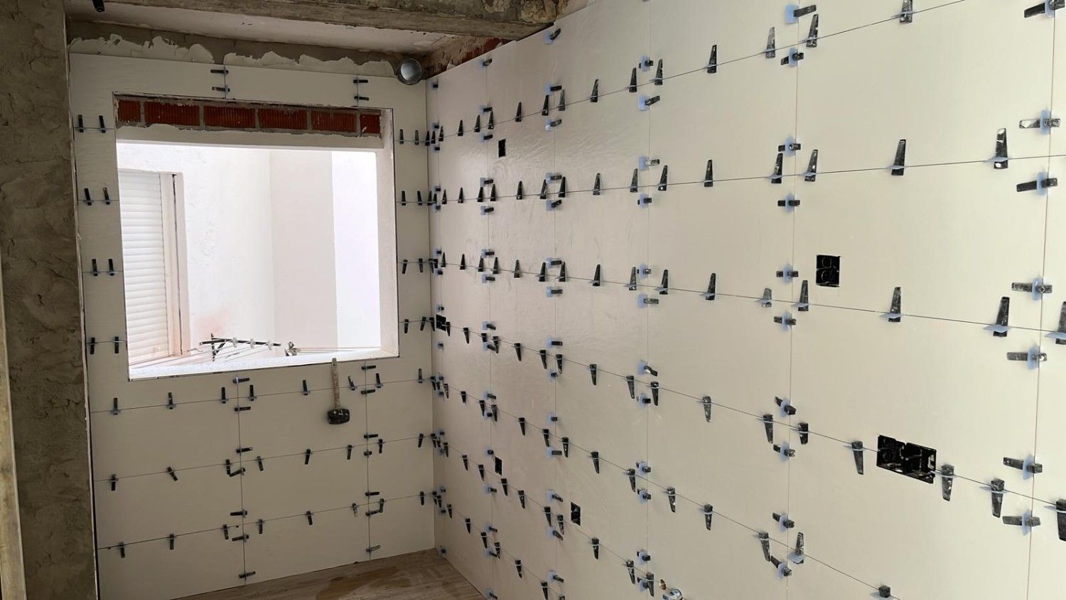 Habitación vacía en proceso de renovación, con paredes blancas y cableado a la vista, vista a través de una ventana.