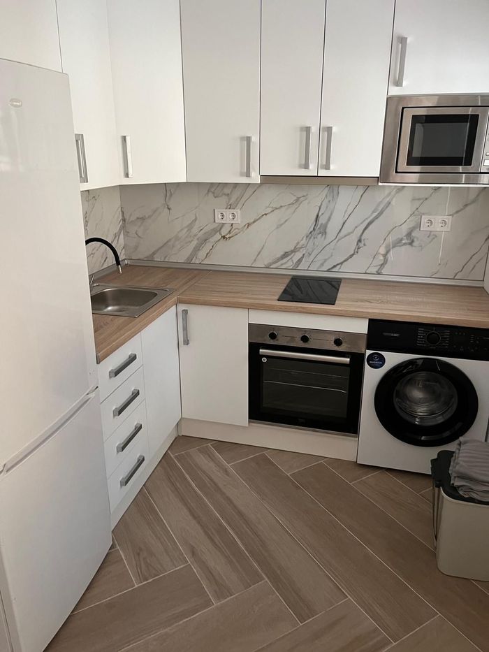 Pequeña cocina moderna con armarios blancos, encimeras de madera, horno empotrado, microondas y lavadora.