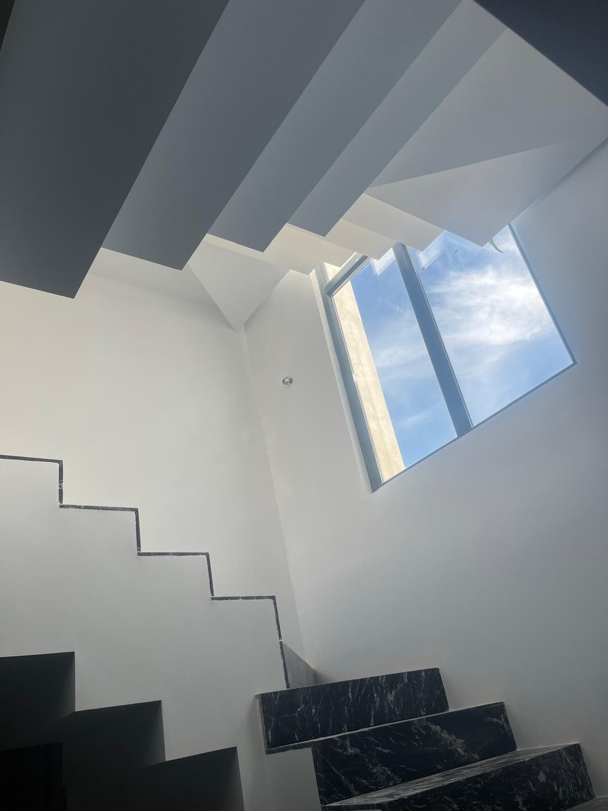 Escalera moderna con paredes blancas, escalones oscuros y una claraboya que deja ver el cielo azul y las nubes.