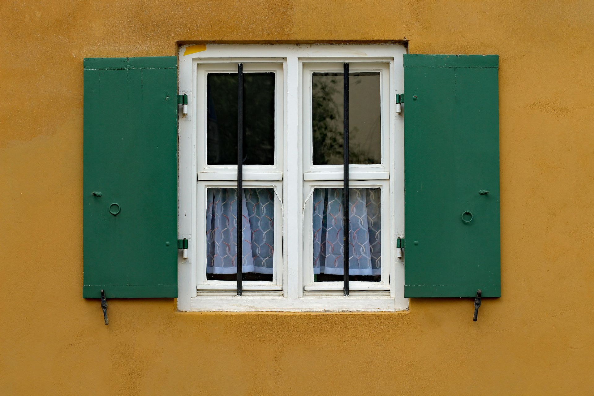 Ventana con contraventanas verdes sobre una pared amarilla, marco blanco, rejas negras y cortinas transparentes.