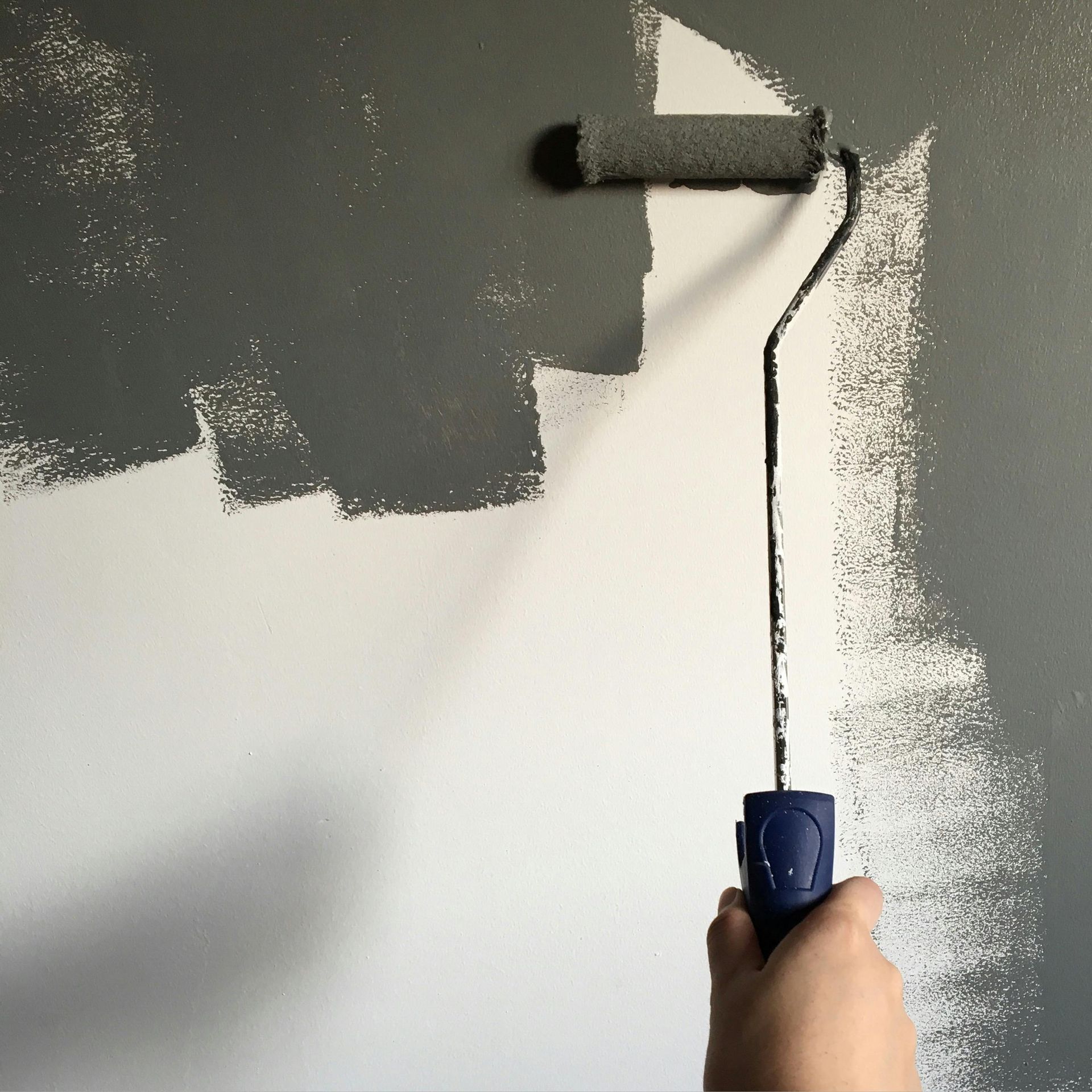 Mano utilizando un rodillo de pintura para aplicar pintura gris sobre una pared blanca.