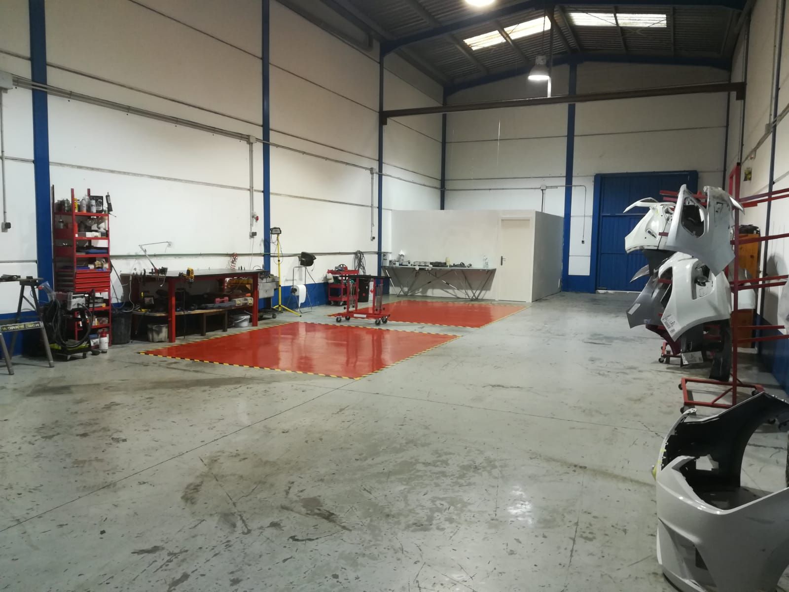 Dentro de un taller con herramientas, suelo rojo y piezas de vehículos. Suelo gris, paredes blancas y una puerta azul.