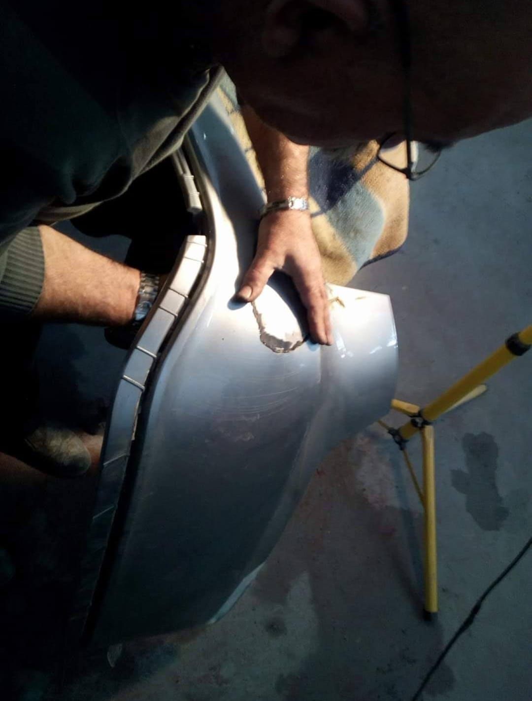 Persona reparando el parachoques de un coche, inspeccionando los daños cerca de una luz.