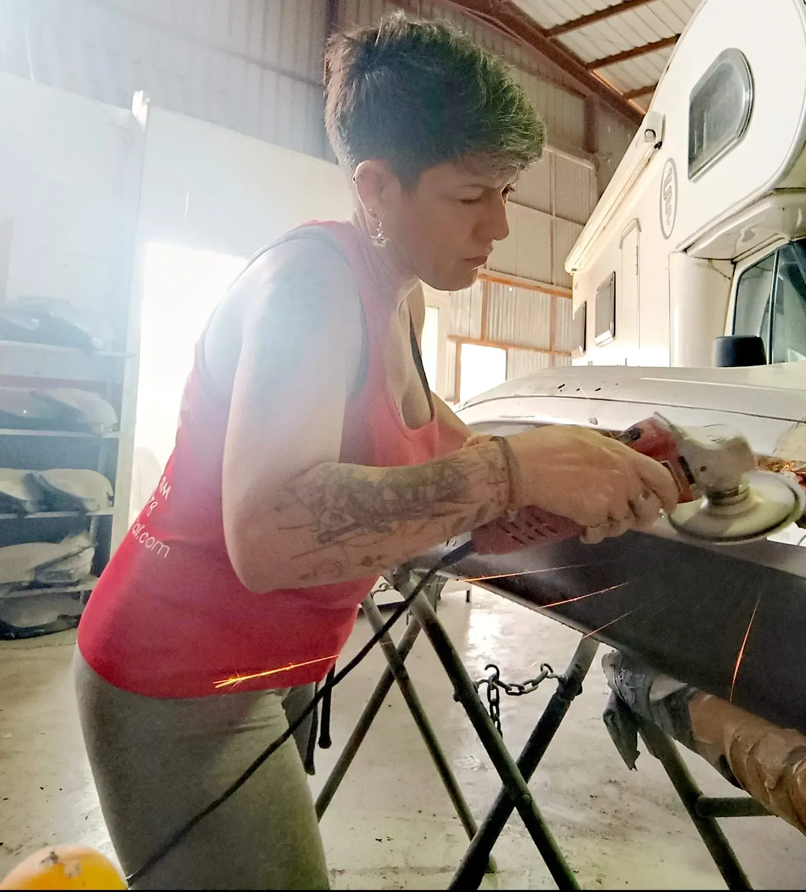 Mujer con cabello teñido de verde moliendo metal en un taller, vestida con una camiseta sin mangas roja y leggings.
