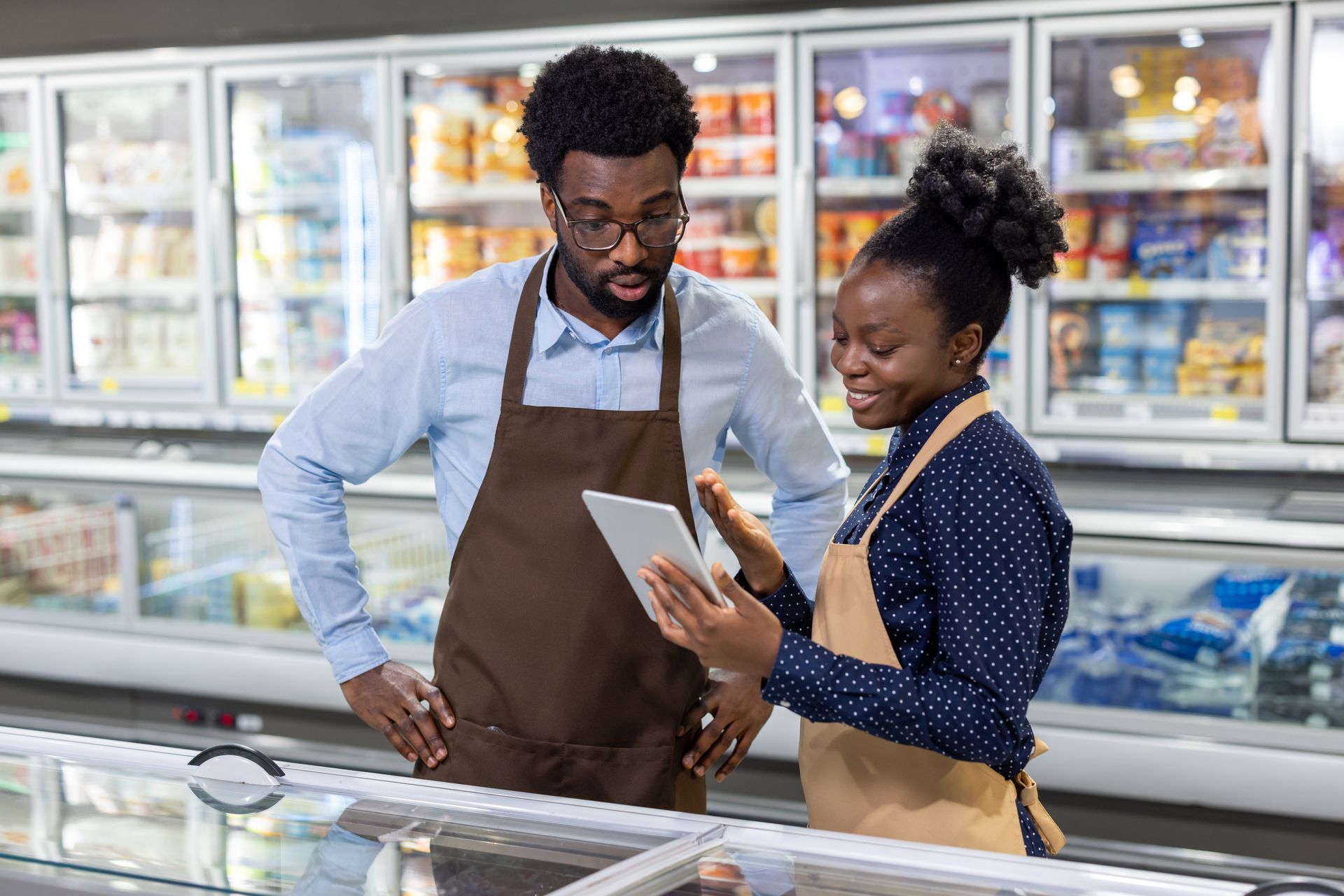 Deux employés de supermarché consultent une tablette à côté d'un rayon de produits surgelés.
