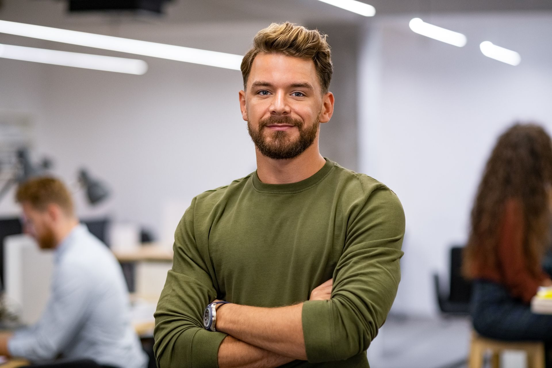 Un homme souriant, vêtu d'une chemise vert olive, se tient debout, les bras croisés, dans un bureau moderne.