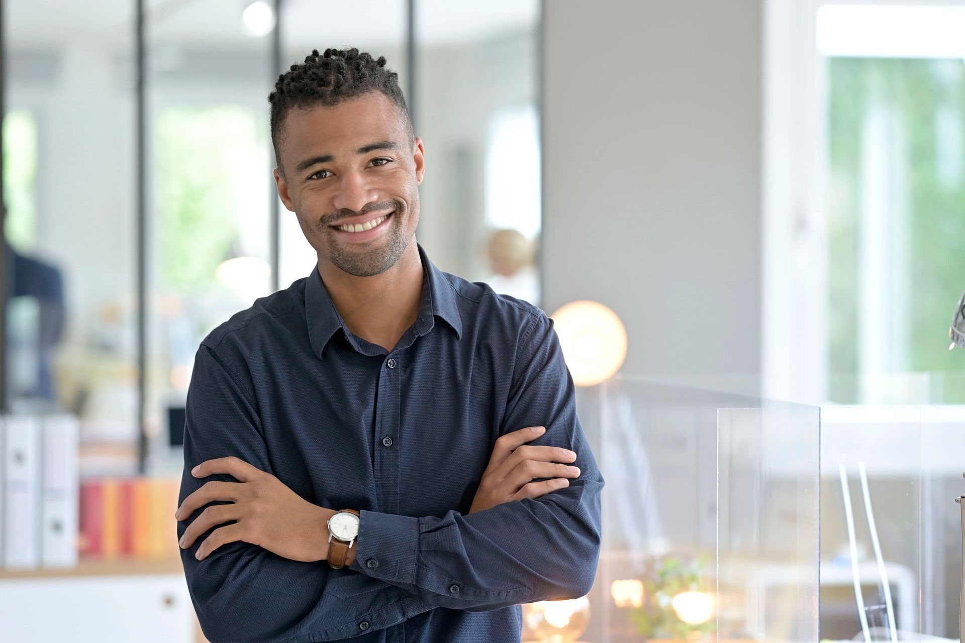 Une personne souriante, vêtue d'une chemise bleu marine, se tient debout, les bras croisés, dans un bureau lumineux.