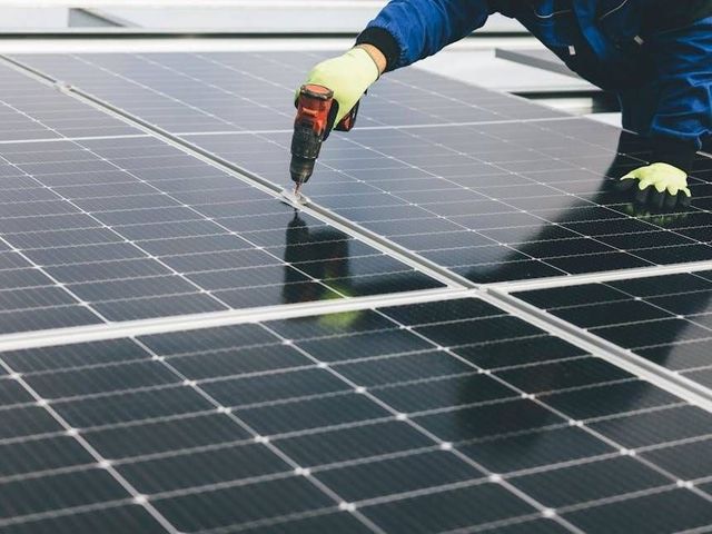 Ein Techniker bohrt Löcher in Solarpaneele auf einem Dach; er trägt Handschuhe und blaue Arbeitskleidung.