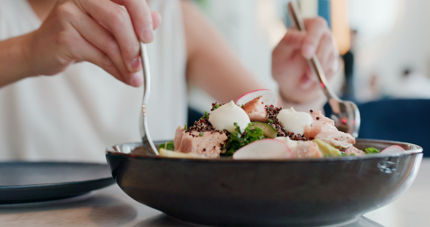 Hände, die mit Besteck einen Salat mit Lachs, Quinoa und Klecksen weisser Sahne aus einer dunklen Schüssel essen.