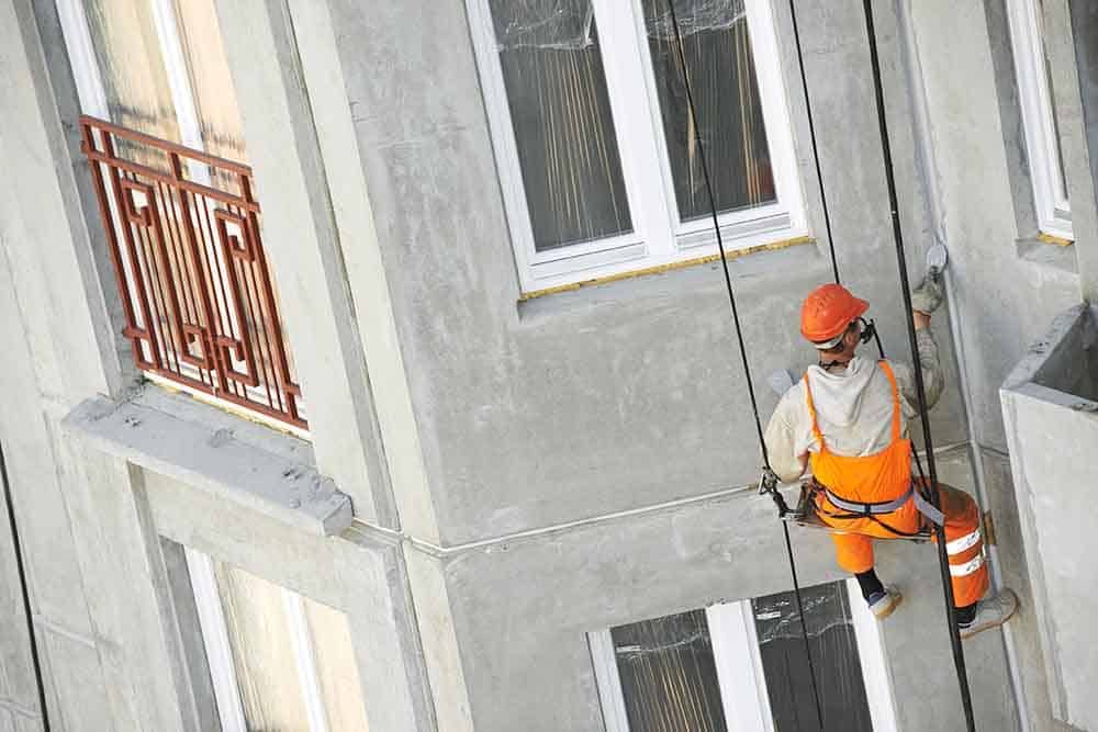Oranssiin turvavarusteisiin pukeutunut työntekijä laskeutuu alas harmaata rakennuksen julkisivua ämpärit vierellään.