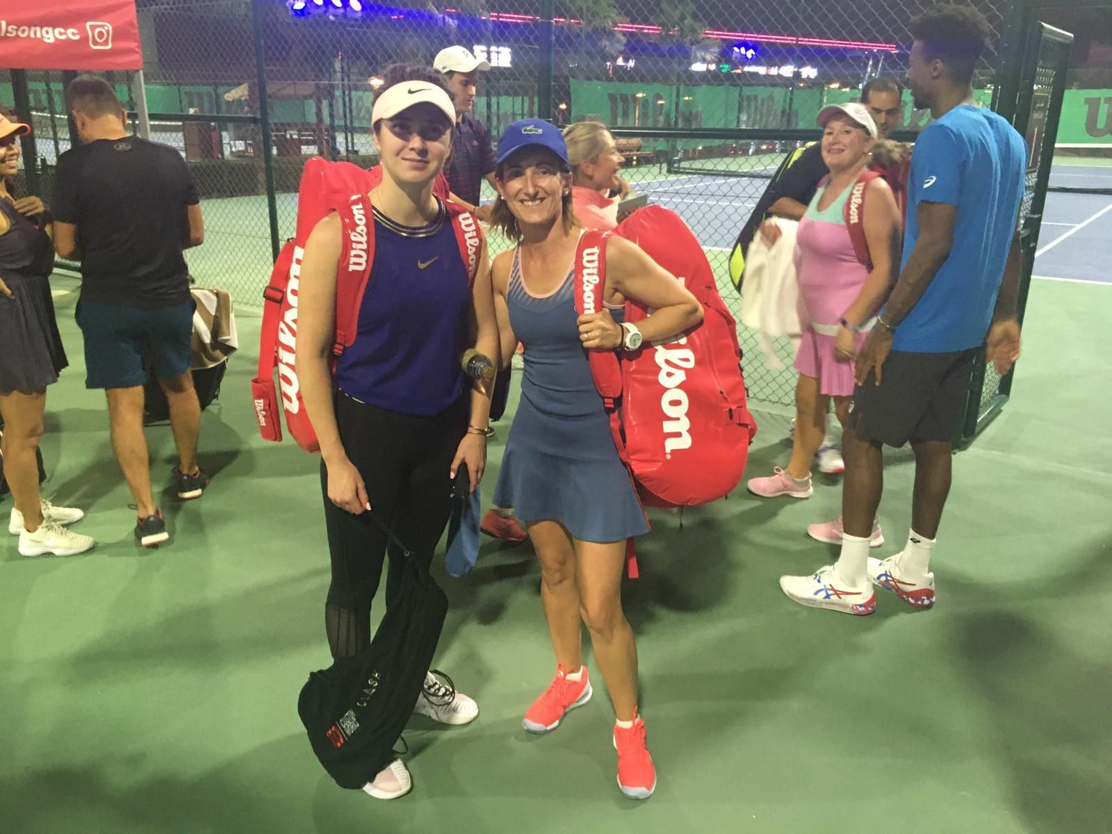 Dos personas posan con bolsas rojas para raquetas de tenis en una cancha cubierta, sonriendo y vestidas con ropa deportiva.