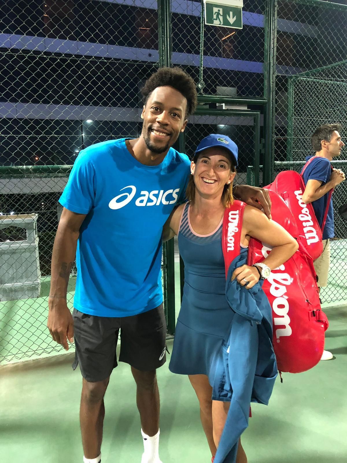 Dos tenistas sonriendo en una cancha, uno sosteniendo una bolsa de raquetas roja y el otro con una camiseta azul de Asics.
