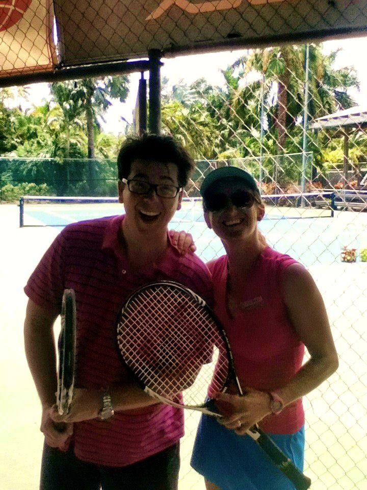 Dos personas con camisetas rosas, sosteniendo raquetas de tenis en una cancha al aire libre, sonríen bajo un toldo que les da sombra.