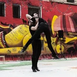 Dos hombres bailan frente a una pared de graffitis.