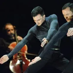 Dos hombres están bailando en un escenario con un violonchelo de fondo.