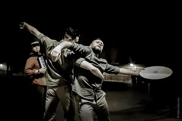 Dos hombres están bailando en una habitación oscura con un hombre tocando un acordeón en el fondo.