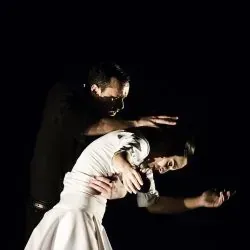 Un hombre sostiene a una mujer con un vestido blanco en una habitación oscura.