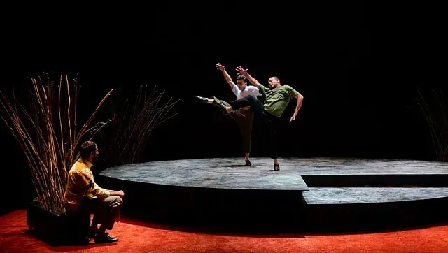 Un hombre está sentado en un escenario mirando a dos bailarines actuar en el escenario.