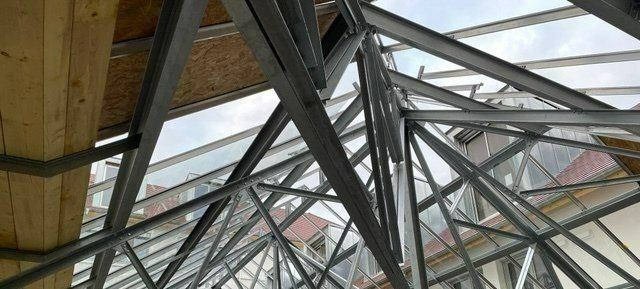 Charpente métallique de toiture avec panneaux de verre, vue de dessous. Poutre de soutien en bois en haut à gauche.