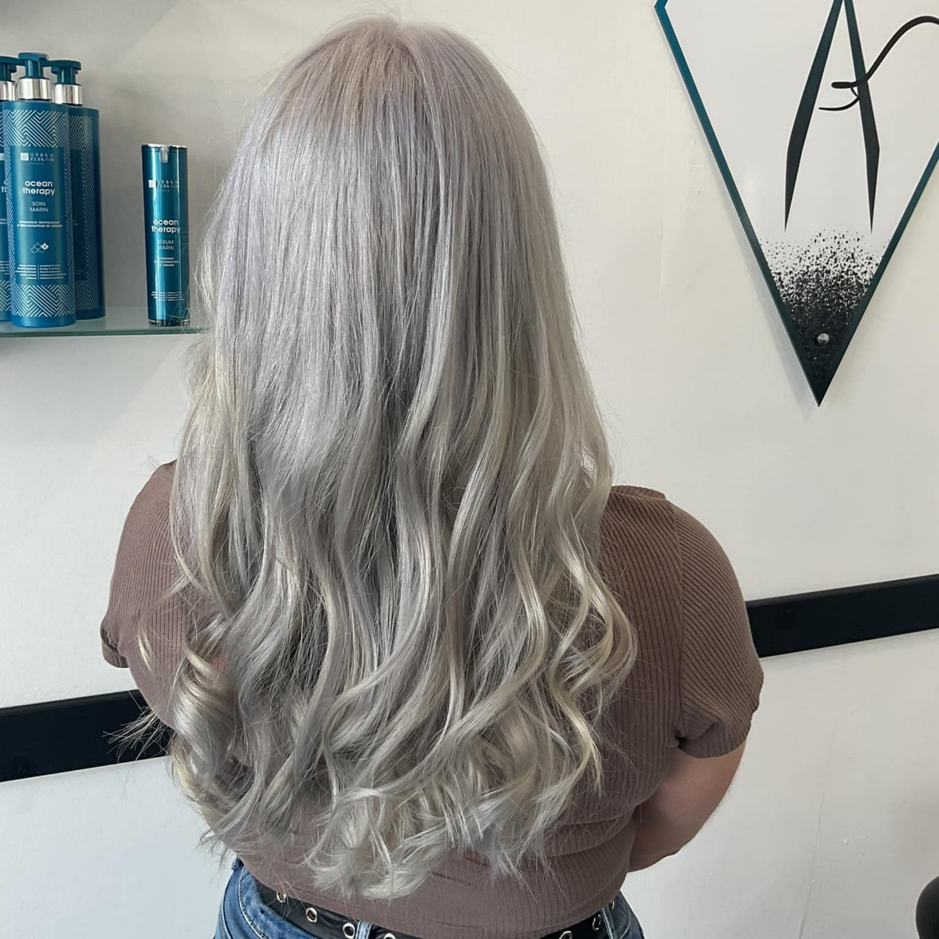 Femme aux longs cheveux ondulés blond argenté. Elle porte une chemise marron dans un salon de coiffure, près des produits.