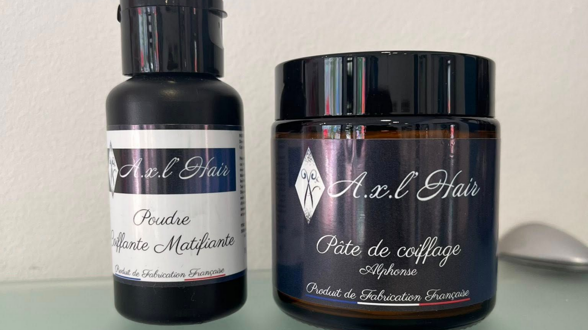 Deux produits A.X.L'Hair : un flacon noir avec une étiquette blanche et un pot avec un couvercle noir.